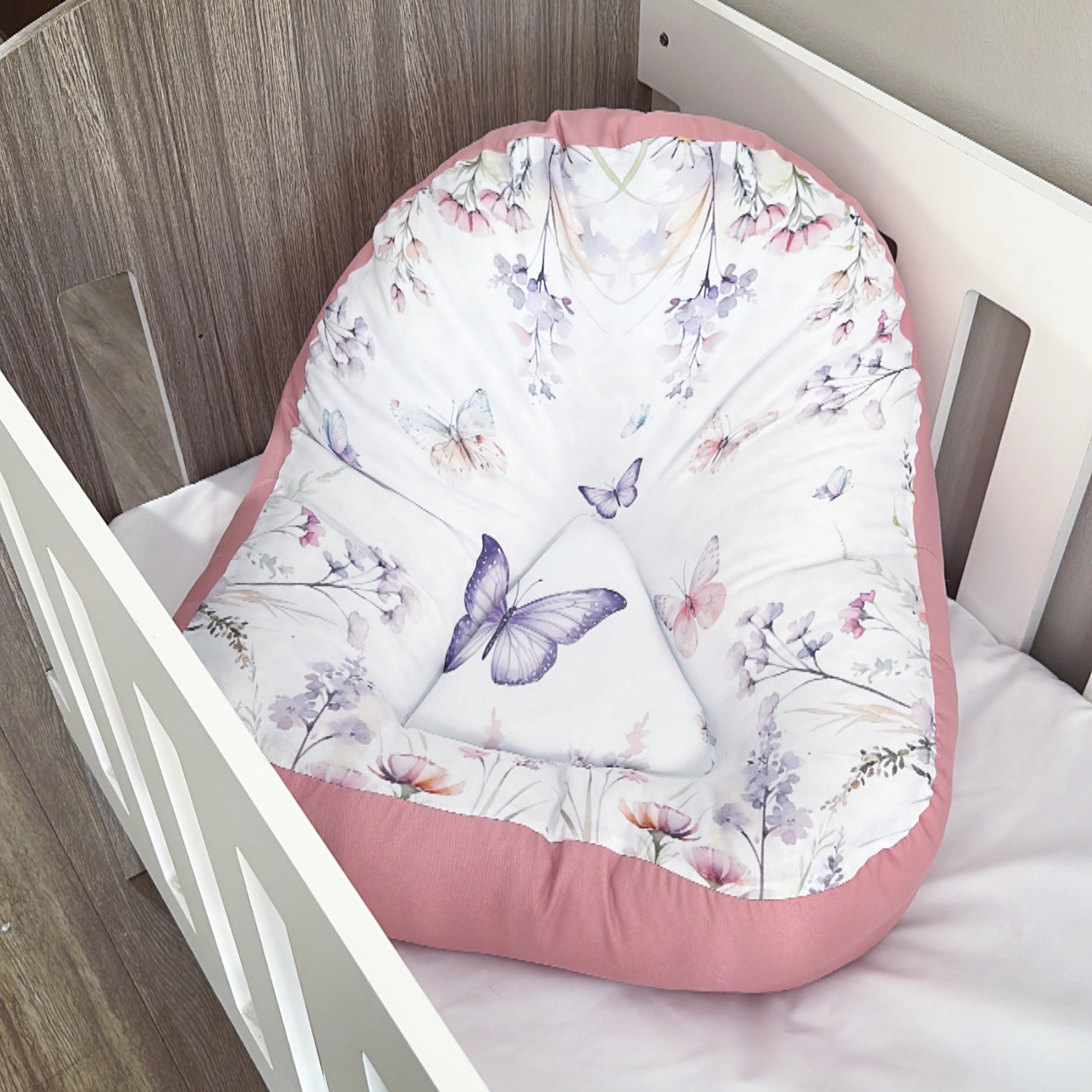 A pink and white floral butterfly patterned lounger for babies, placed inside a crib.