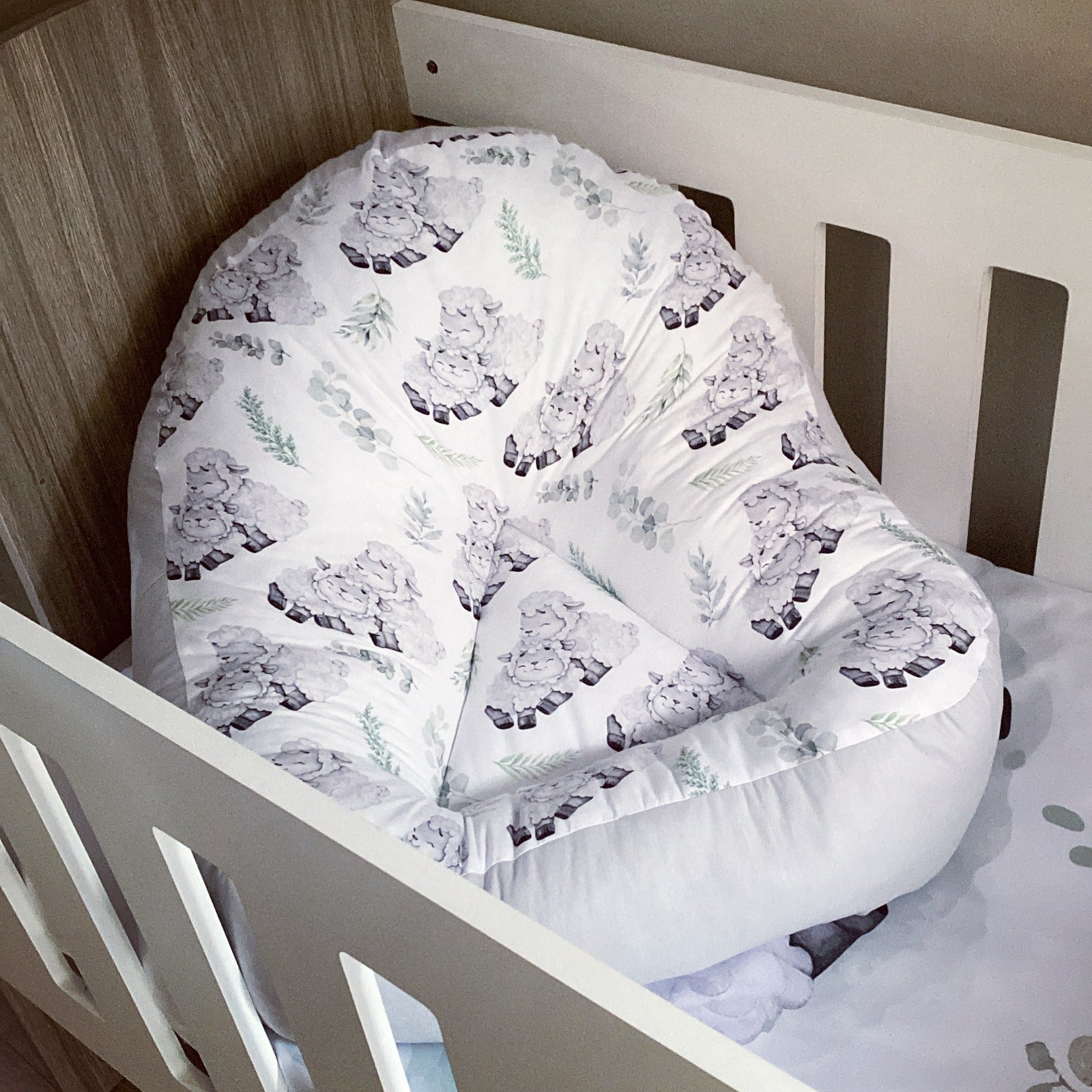 A white fabric lounger with a sheep pattern, positioned in a wooden crib.